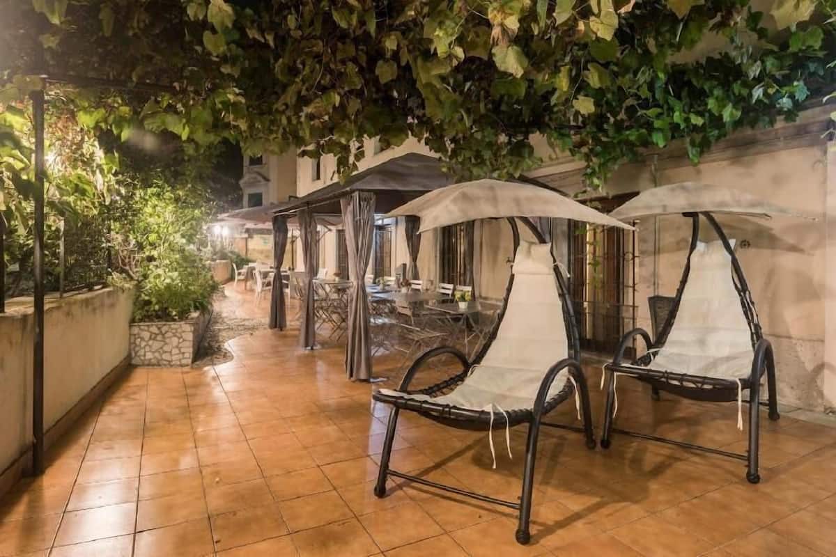 An outdoor patio with metal chairs and umbrellas, surrounded by greenery and a tiled floor.