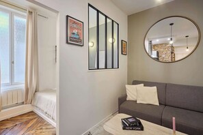Ein modernes Wohnzimmer mit einem grauen Sofa, einem kleinen Couchtisch mit Büchern, einem Spiegel und einem gerahmten Bild an der Wand.