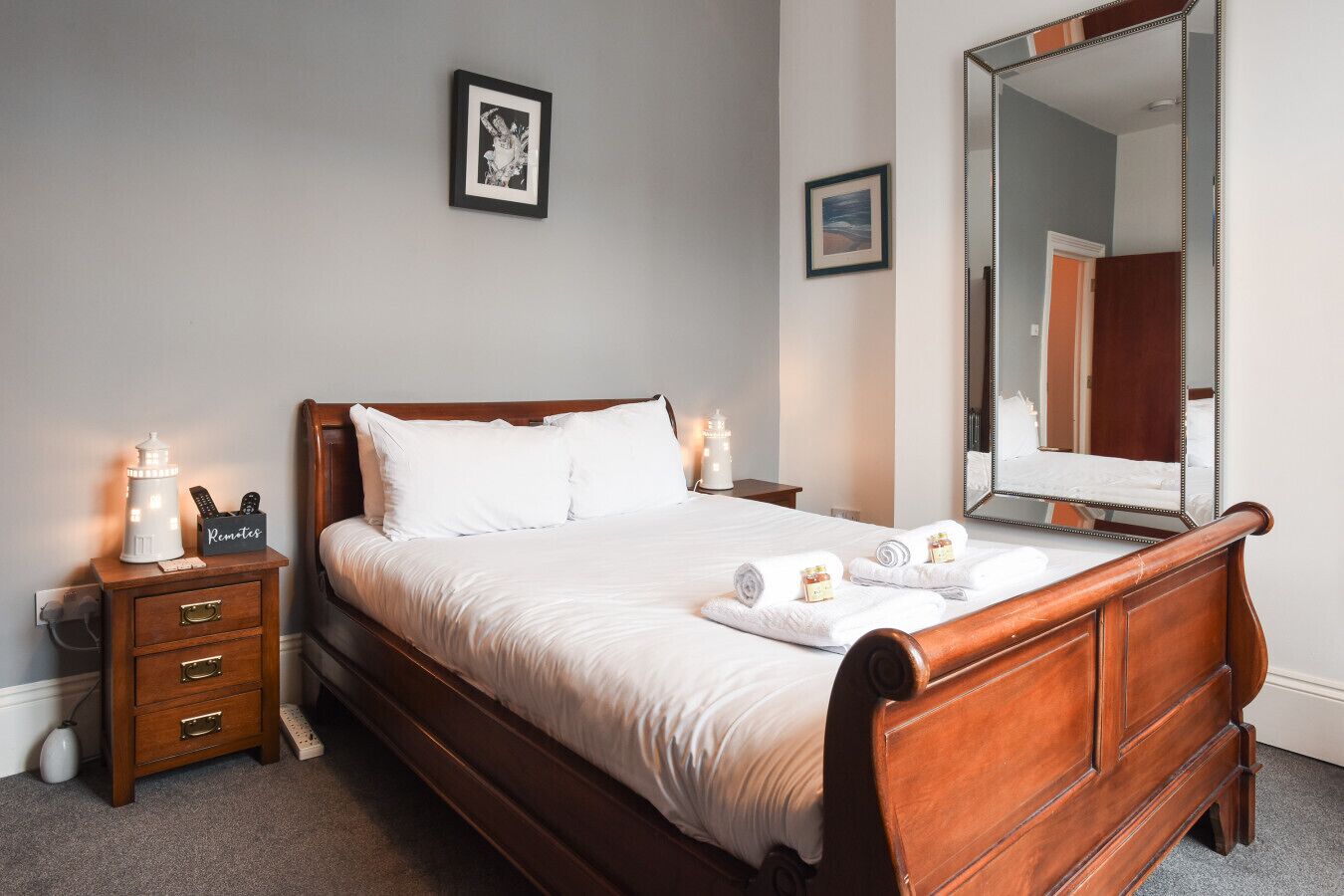 A neatly made bed with white linens, a bedside lamp, a remote control holder, and a small nightstand with drawers.