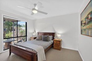 A bedroom with a bed, bedside tables, a ceiling fan, and a large window. - Swell Beach House - Families Walk to the Beach (Second Valley)
