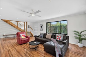 A spacious living area with a staircase, leather sofas, a coffee table, and a large window offering a view of greenery. - Swell Beach House - Families Walk to the Beach (Second Valley)