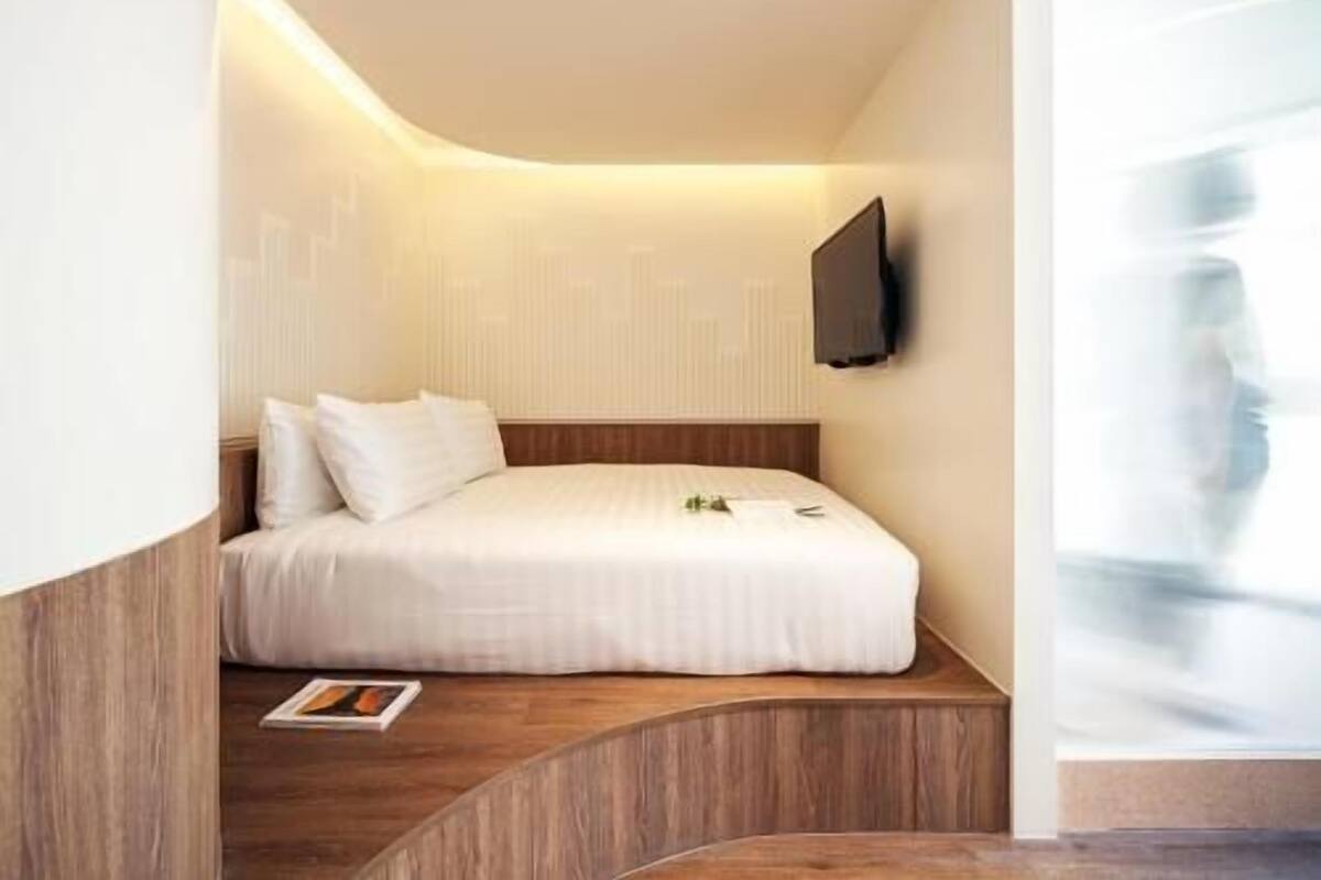 A modern hotel room with a curved wooden platform, a bed with white bedding, a flat-screen TV mounted on the wall, and a magazine on the platform.