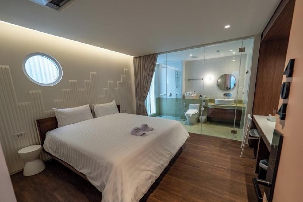 Una habitación de hotel moderna con una cama grande, un ventanal redondo, un baño con paredes de vidrio y suelo de madera.