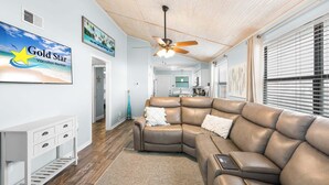 A living room with a brown leather sectional sofa, a white console table, a ceiling fan, and a large window with blinds. - Lani Kai 230 Gulf Coast Condo. Walk to Beach + Pools + Balcony Views! (Gulf Shores)