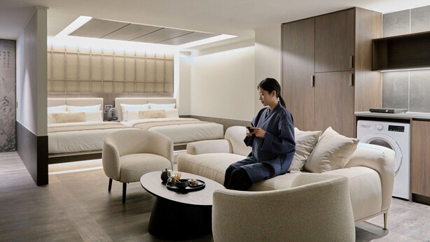 Quarto de hotel moderno com sofá, mesa de centro, cama com encosto, máquina de lavar e uma janela grande.