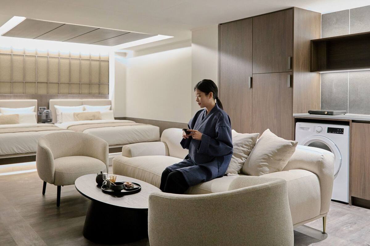 A modern hotel room with a sofa, a coffee table, a bed with a headboard, a washing machine, and a large window.