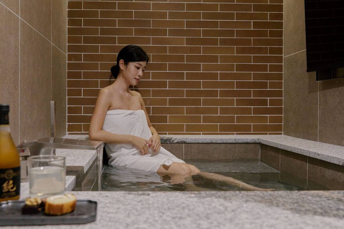 A person in a spa bath with a towel, a bottle, and a glass on the counter.