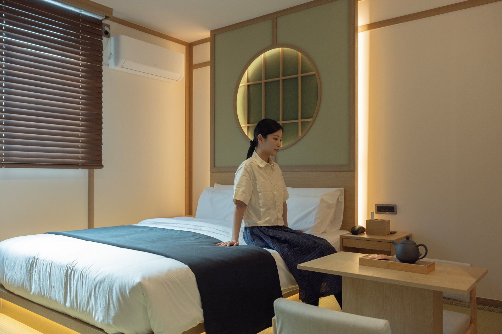 A hotel room with a bed, a desk with a teapot, and a wall-mounted air conditioner.