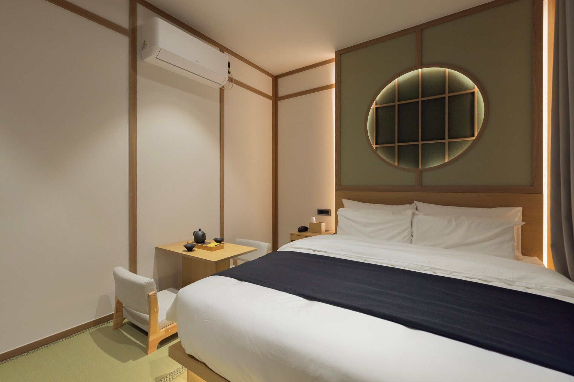 A hotel room with a bed, a chair, a small table, and a wall-mounted air conditioner.