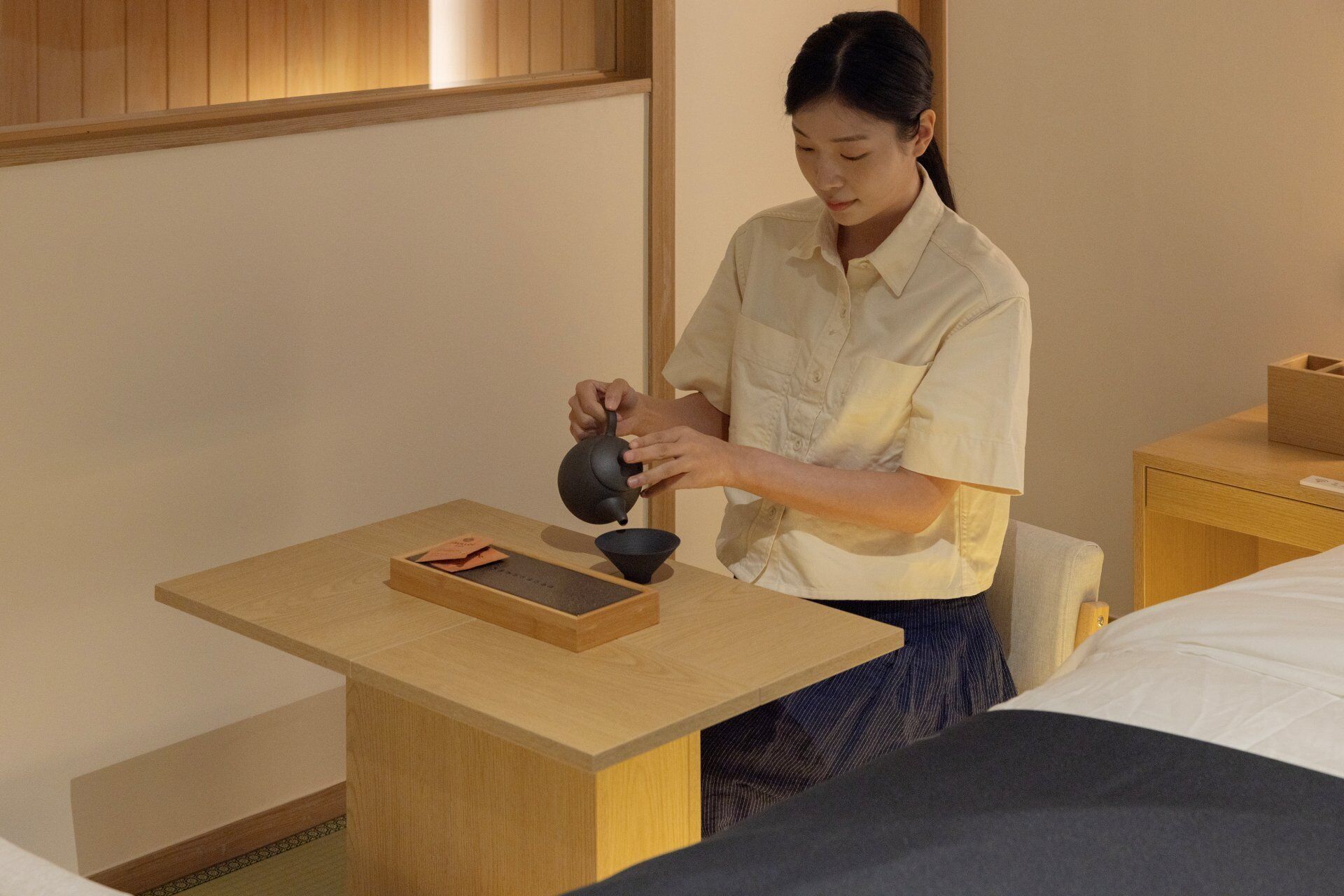 Una persona sirve té en una taza sobre una mesa de madera en una habitación con una cama y una mesita de noche.