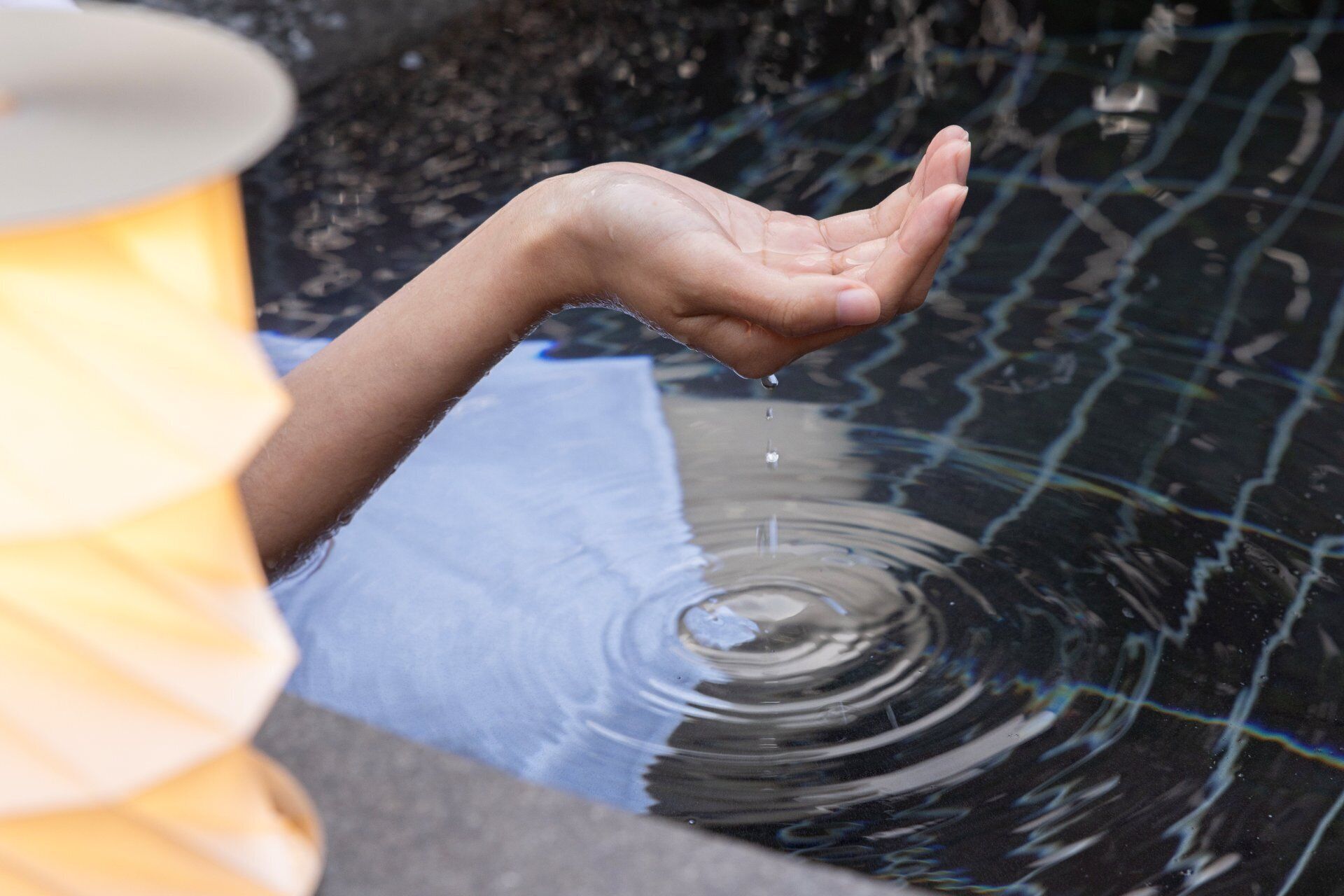 A hand reaching into a body of water, possibly a pool or fountain, with a blurred background.