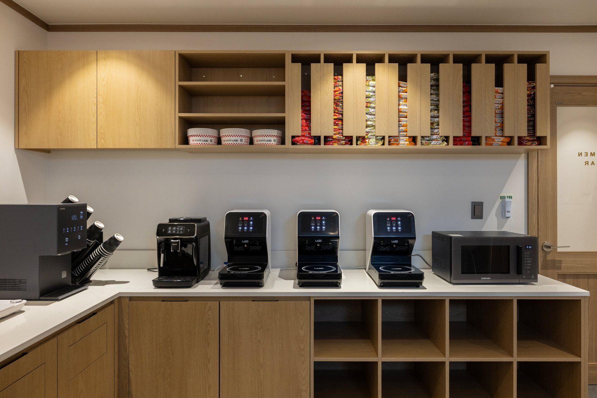 A kitchen with five coffee machines, a microwave, and shelves stocked with snacks.