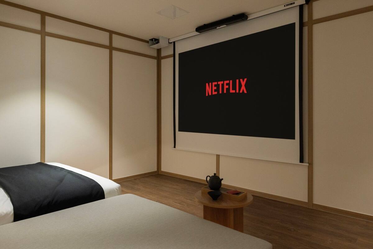 A room with a bed, a small table with a teapot, and a wall-mounted screen displaying 'NETFLIX'.