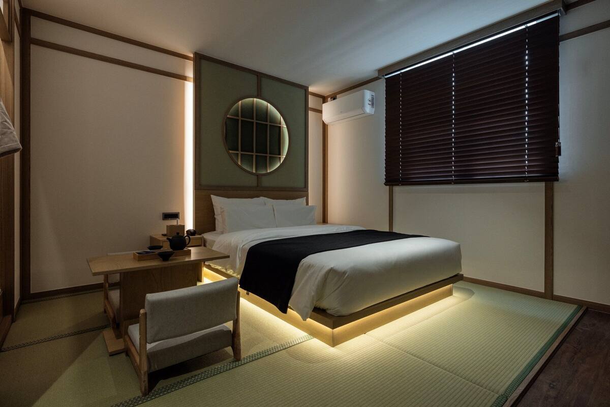 A hotel room with a bed, bedside table, chair, and a window with blinds.