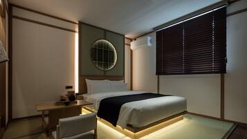 A hotel room with a bed, bedside table, chair, and a window with blinds.