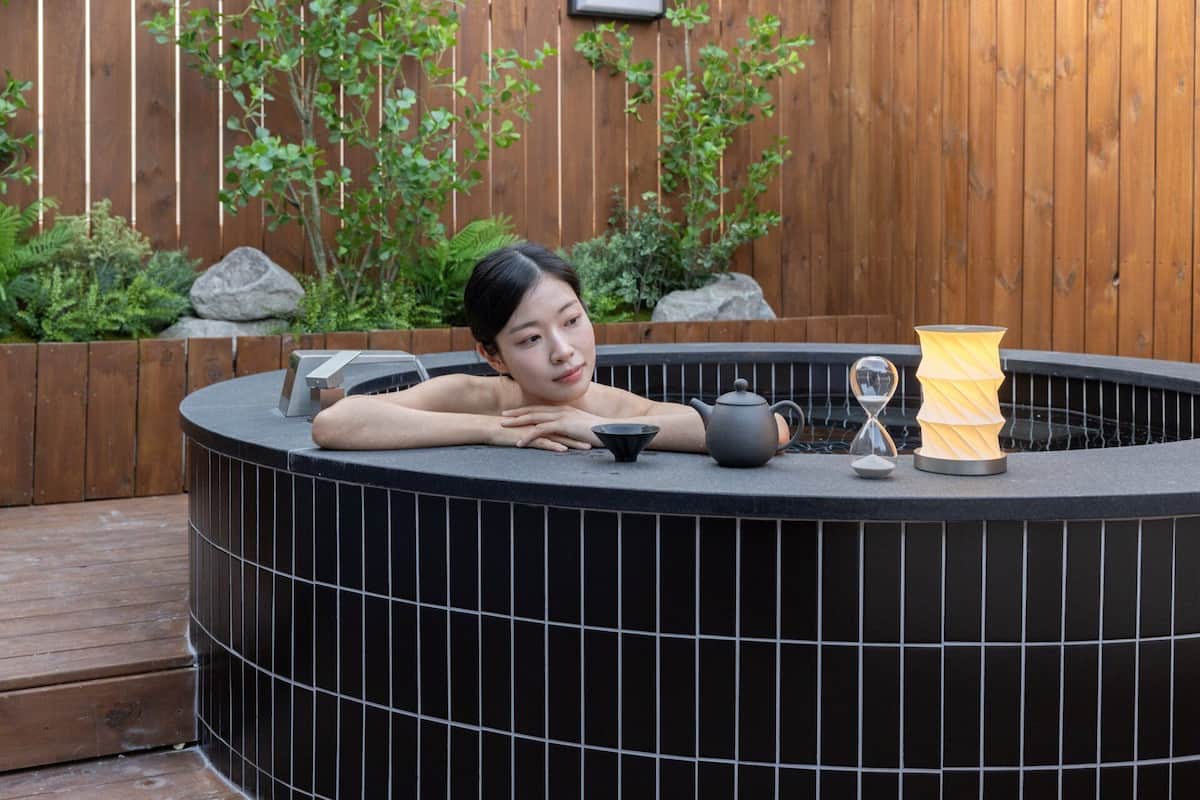 A person relaxing in a black outdoor hot tub with a teapot, a small bowl, and an hourglass.