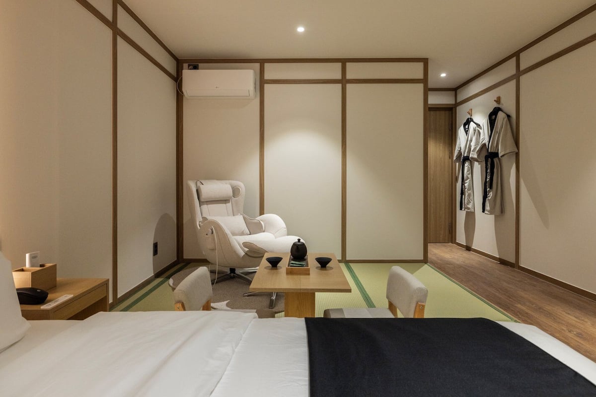 A hotel room with a bed, a massage chair, a table with a teapot, and a wall-mounted robe rack.