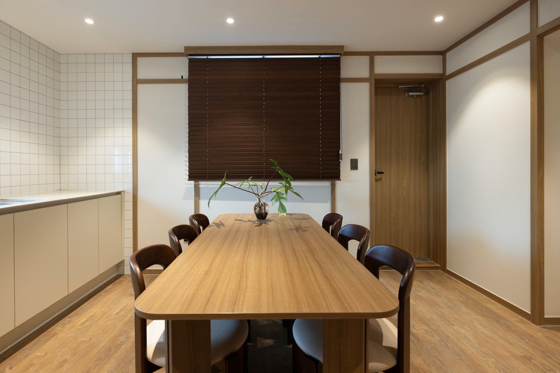 A conference room with a wooden table, chairs, a potted plant, and a window with blinds.