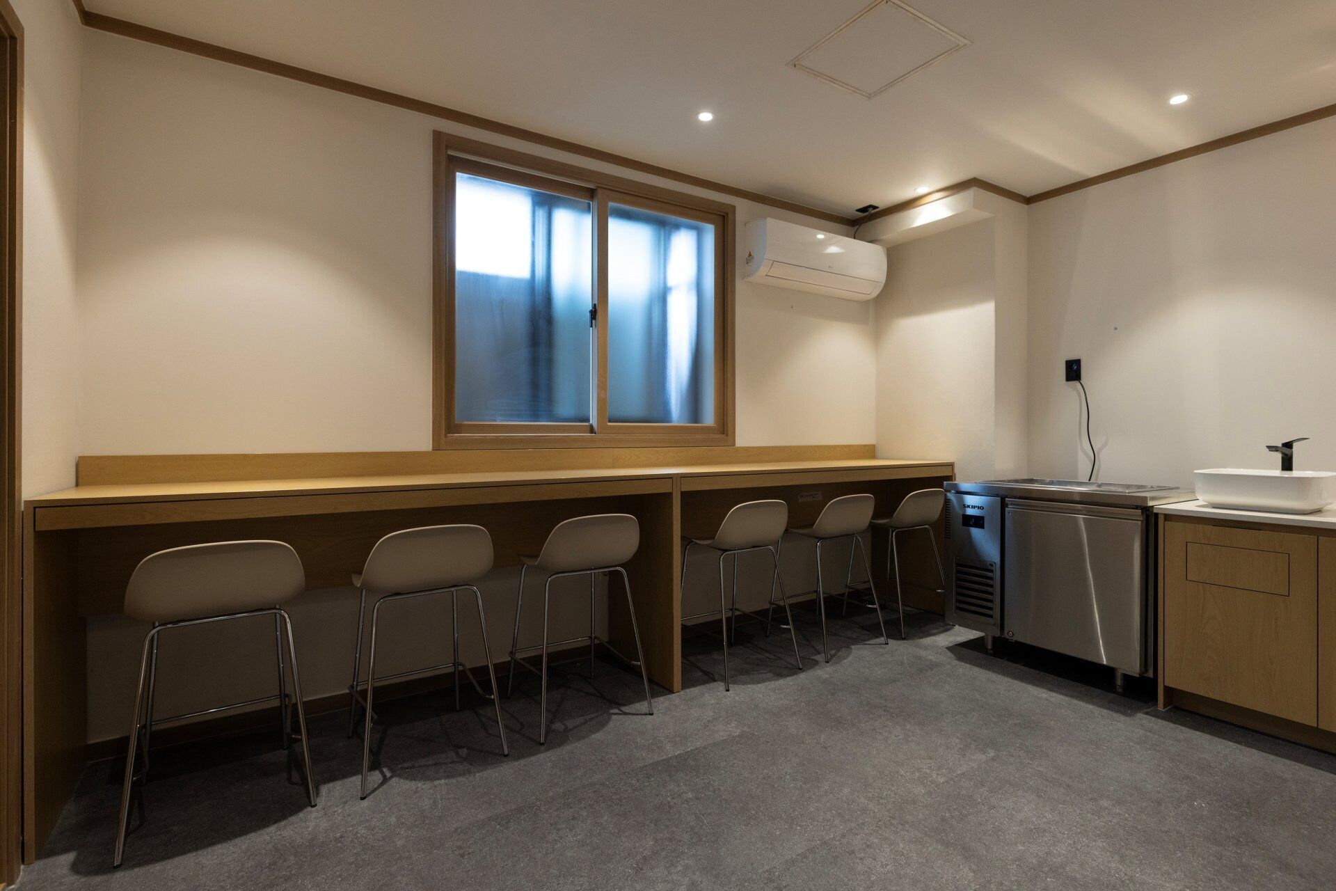Uma sala de conferências com uma mesa comprida, cadeiras, uma janela, unidade de ar condicionado, pia e frigorífico.