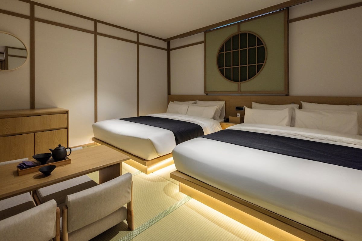 A hotel room with two beds, a desk with a teapot, a mirror, and a window with a grid pattern.