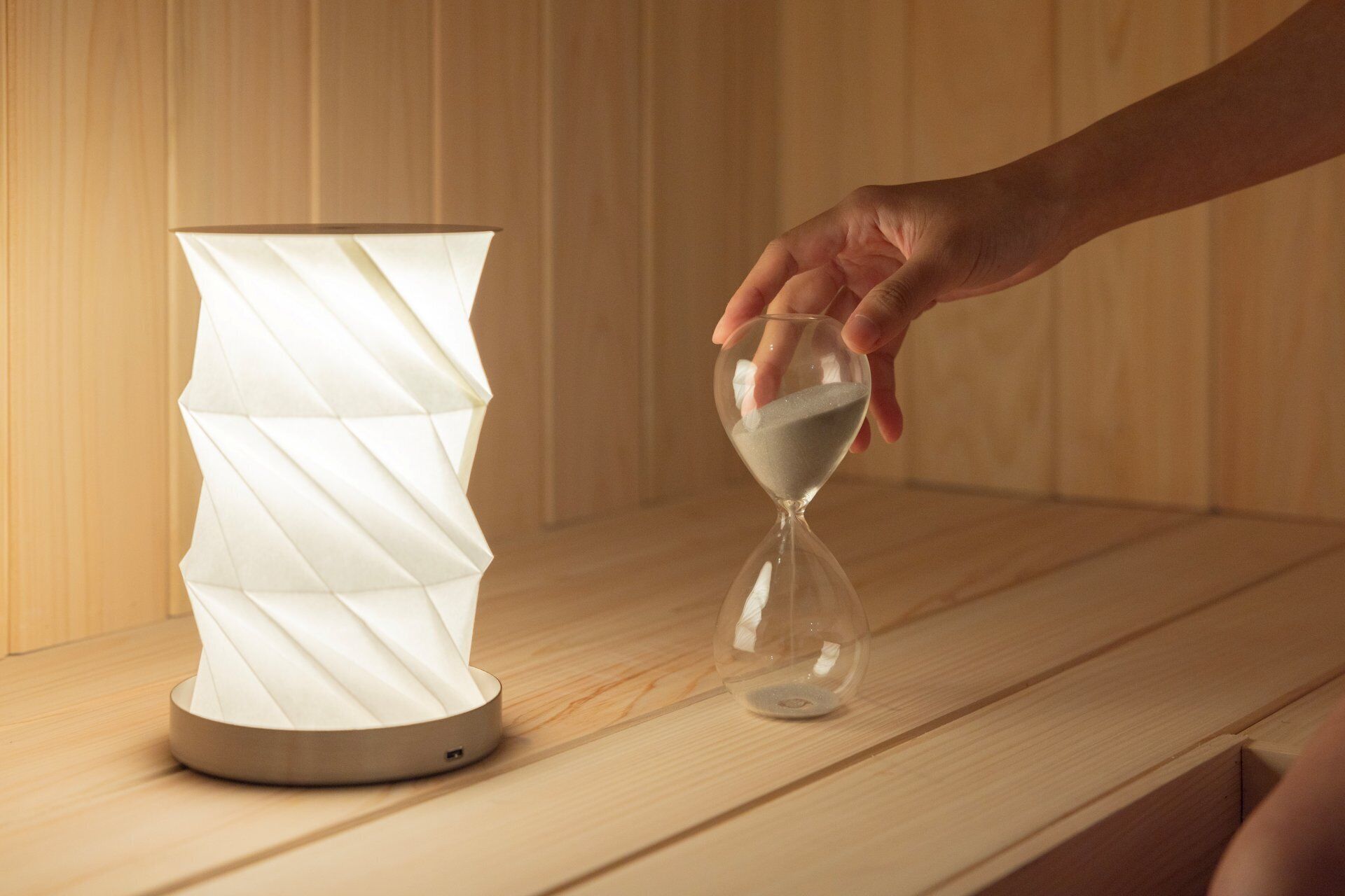 Une main tenant un sablier à côté d’une lampe moderne, le tout posé sur une surface en bois.