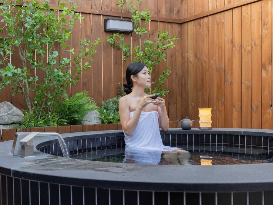 A person in a hot tub with a wooden fence and greenery in the background.