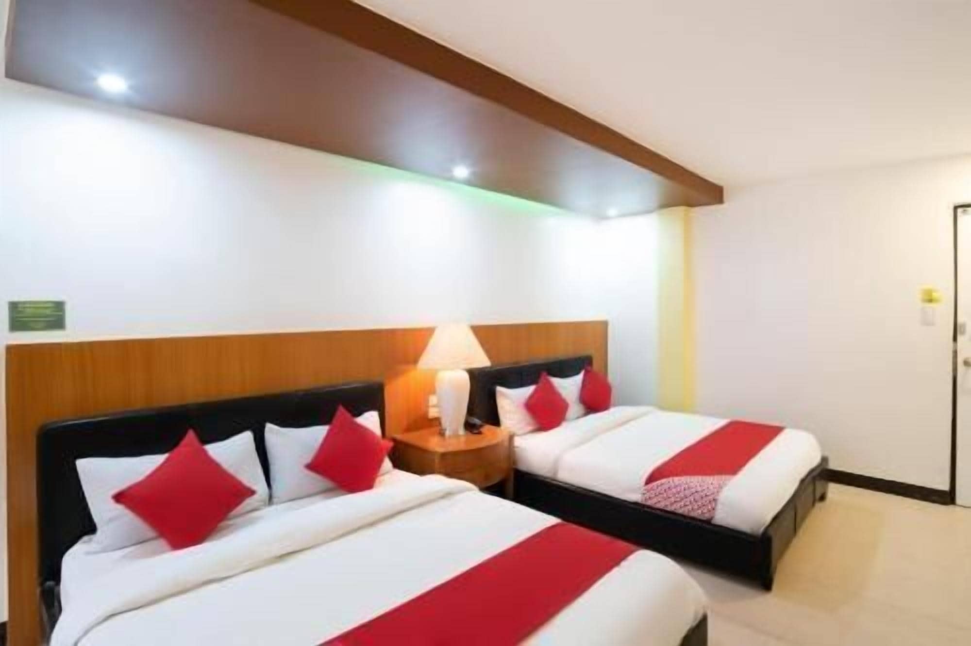 A hotel room with two beds, each with red pillows and a white bedspread. There is a wooden headboard, a bedside table with a lamp, and a door to the right.