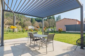 Eine überdachte Terrasse mit Tisch und Stühlen, ein Metallstuhl und Blick auf eine grüne Rasenfläche sowie Gebäude im Hintergrund.