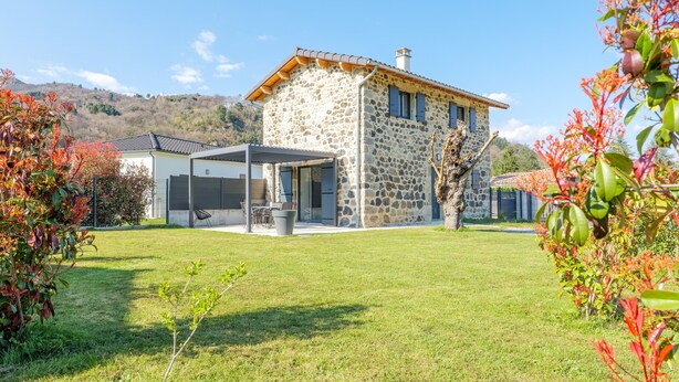 Ein Steinhäuschen mit überdachter Terrasse, einem Rasen und einem Berg im Hintergrund.