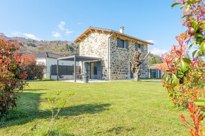Une maison en pierre avec un patio couvert, une pelouse et une montagne en arrière-plan.