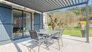Eine Terrasse im Freien mit Tisch und Stühlen, einem überdachten Bereich und Blick auf einen bewaldeten Hang.