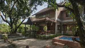 A two-story house with a covered porch, a swimming pool, and a garden with various plants and trees. - Amã Stays & Trails Asanji Wadi (Alibag)