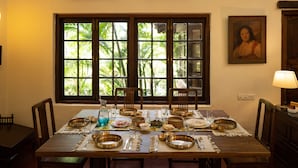 A dining table set with traditional dishes, chairs, and a window with a view of greenery. - Amã Stays & Trails Asanji Wadi (Alibag)