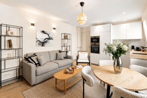 Een moderne woonkamer met een bank, salontafel en stoelen, een eettafel met een vaas, en een keuken met kasten en apparatuur.