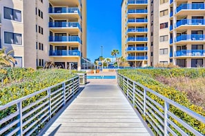 A wooden boardwalk leading to a pool area surrounded by lush greenery and high-rise buildings with balconies. - Four Seasons 701W | 720' Fishing Pier | Beachfront | My Beach Getaways (Orange Beach)