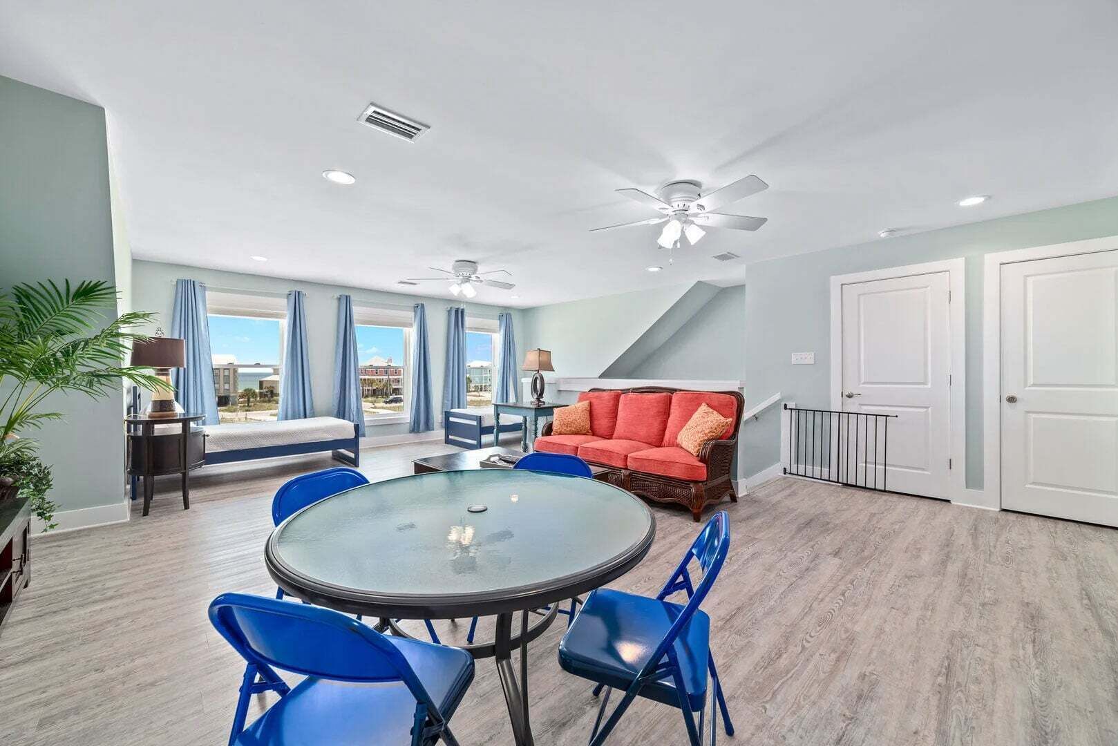 A room with a round glass table, blue chairs, a red sofa, a ceiling fan, and a view of the outside.