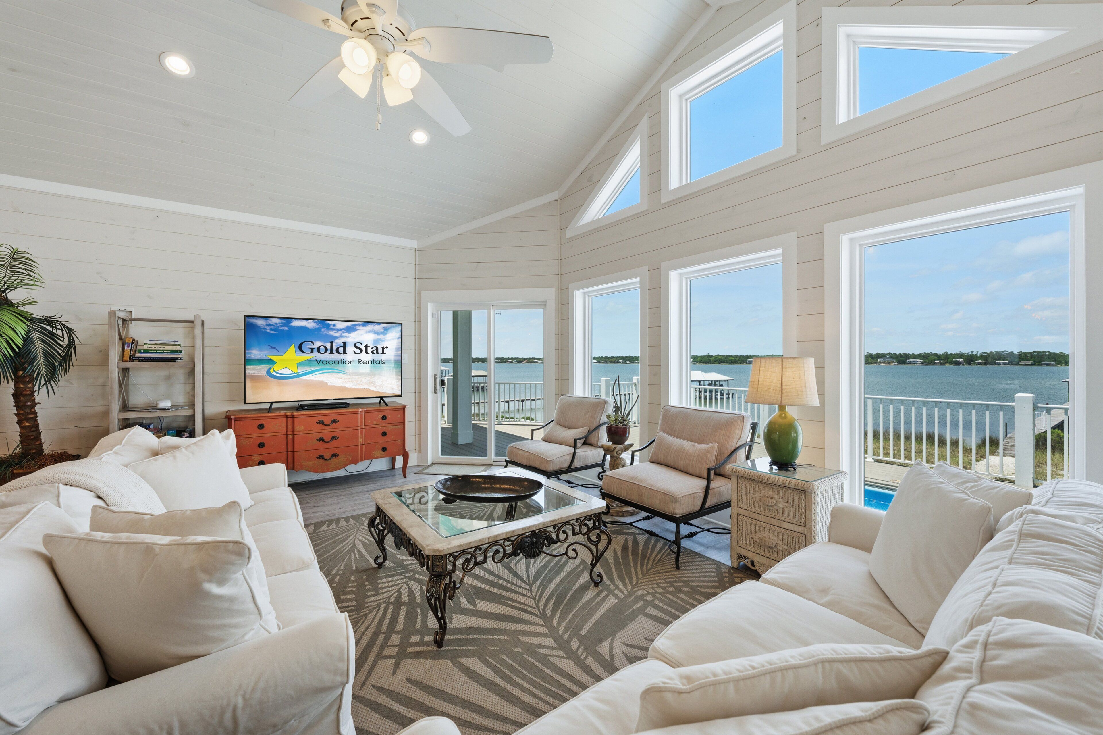 A spacious living room with a large TV, comfortable seating, a ceiling fan, and a view of the ocean.