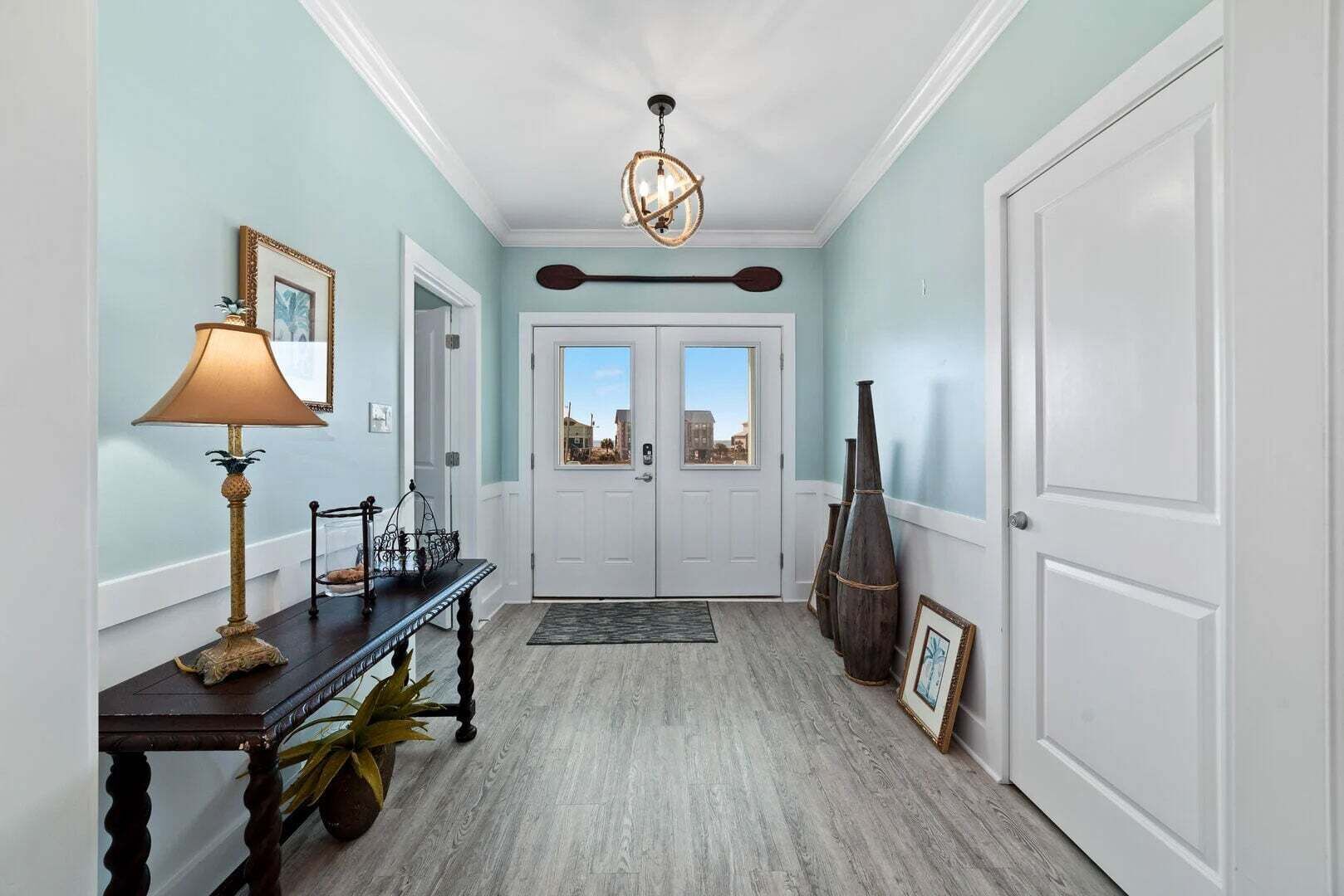 A hallway with a wooden floor, a lamp, a table with a plant, and a door with a view.