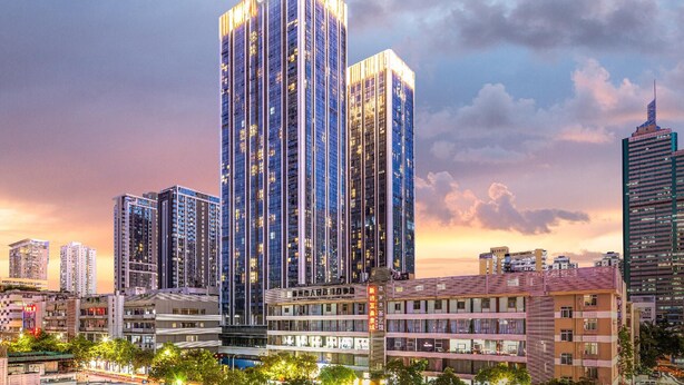 Un edificio alto con varios pisos, rodeado de otros edificios y árboles.