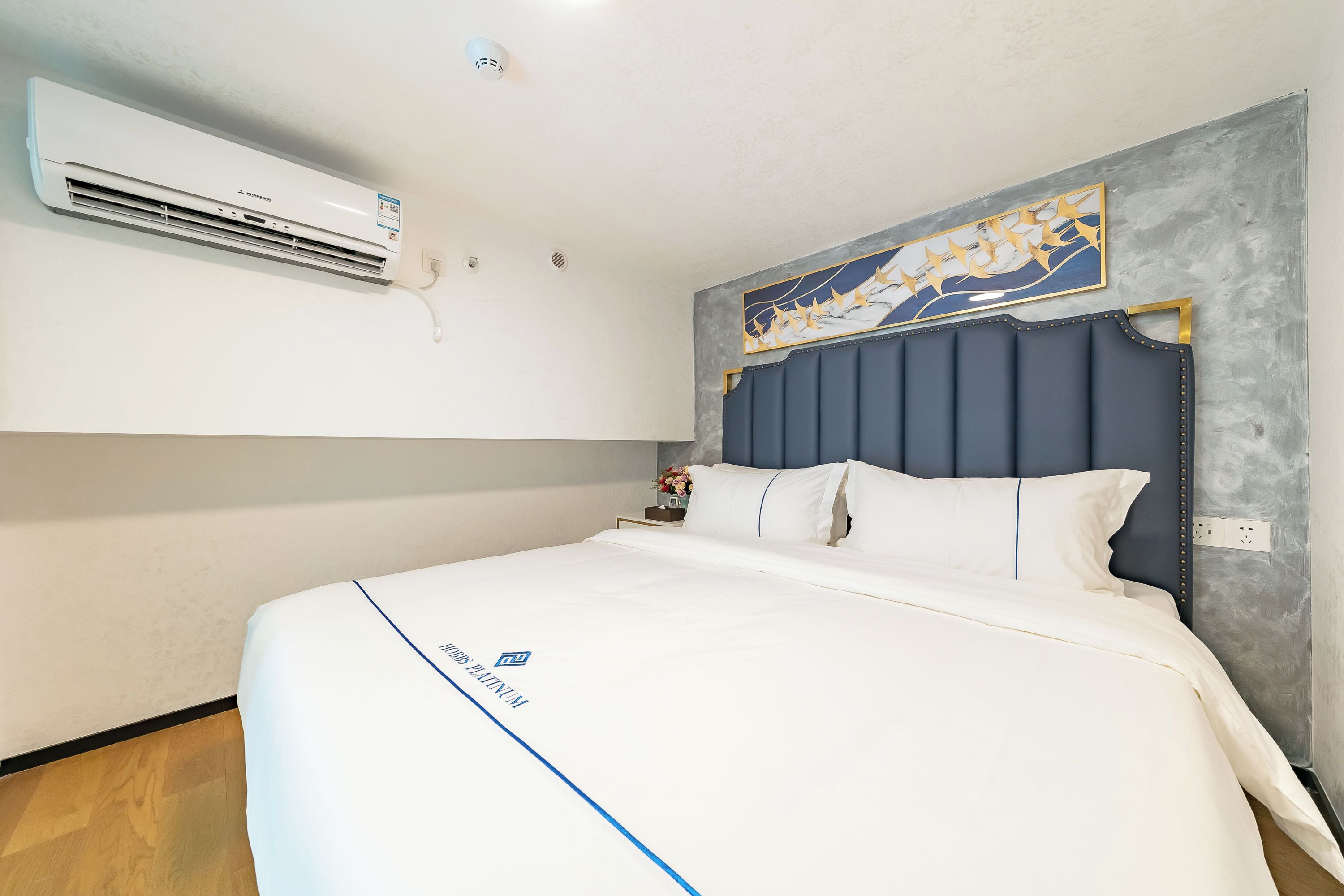 A hotel room with a bed, a wall-mounted air conditioner, a decorative painting, and a nightstand with a vase.