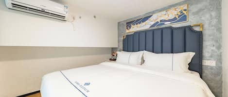 A hotel room with a bed, a wall-mounted air conditioner, a decorative painting, and a nightstand with a vase.