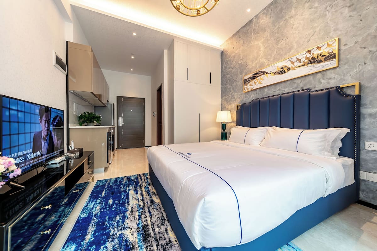 A modern hotel room with a large bed, a television, a desk, and a kitchen area.