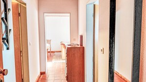 Een gang met een houten plafond, een betegelde vloer en een deur die leidt naar een kamer met een stoel en een kleine tafel.
