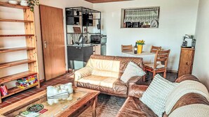 Een woonkamer met een bruine leren bank, een houten salontafel, een boekenkast en een eethoek met een tafel en stoelen.