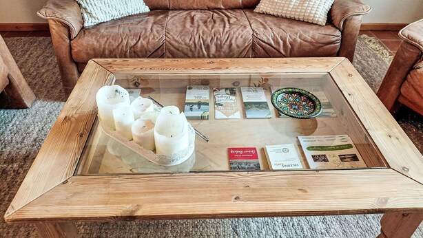 Een bruine leren bank met witte en beige kussens, een glazen salontafel met boeken en kaarsen, en een houten bijzettafeltje.
