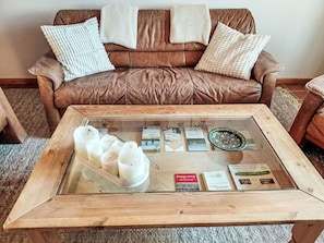 A brown leather sofa with white and beige cushions, a glass coffee table with books and candles, and a wooden side table. - Nice apartment for 2 guests with WIFI, TV, patio and pets allowed (Schieder-schwalenberg)