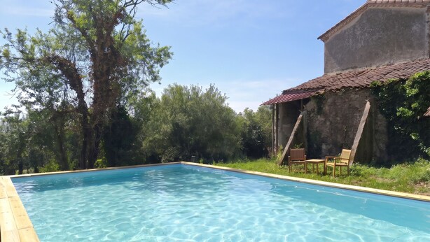 Une piscine aux eaux cristallines, bordée d’une terrasse en bois, entourée de verdure et d’un bâtiment en pierre doté d’un toit en tuiles.