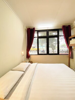 A hotel room with a bed, a nightstand, a window with curtains, and a view of the street outside. - Ars Inn Hotel Yogyakarta (Sewon)