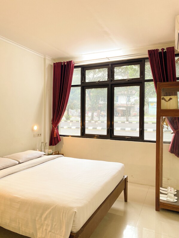 A hotel room with a large bed, a wooden nightstand, a mirror, and a window with curtains. - Ars Inn Hotel Yogyakarta (Sewon)