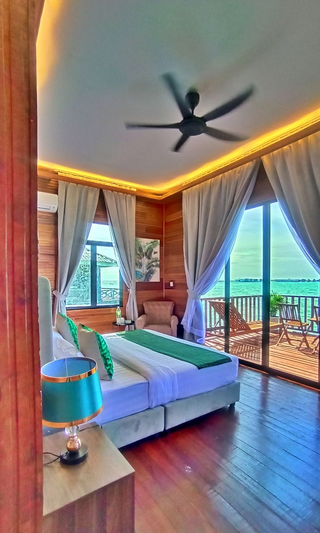 A room with a bed, a ceiling fan, a window with curtains, and a view of the sea.